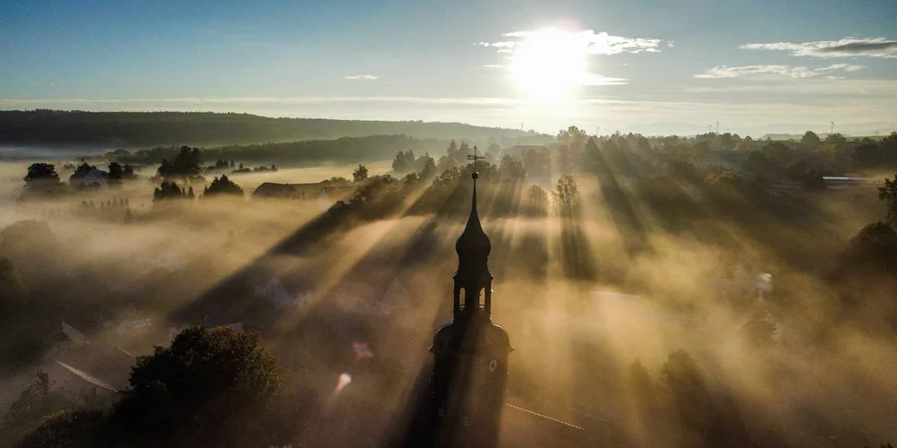 Luftaufnahme von Schönau-Berzdorf mit Nebel und Sonnenstrahlen, im Vordergrund ein Kirchturm.